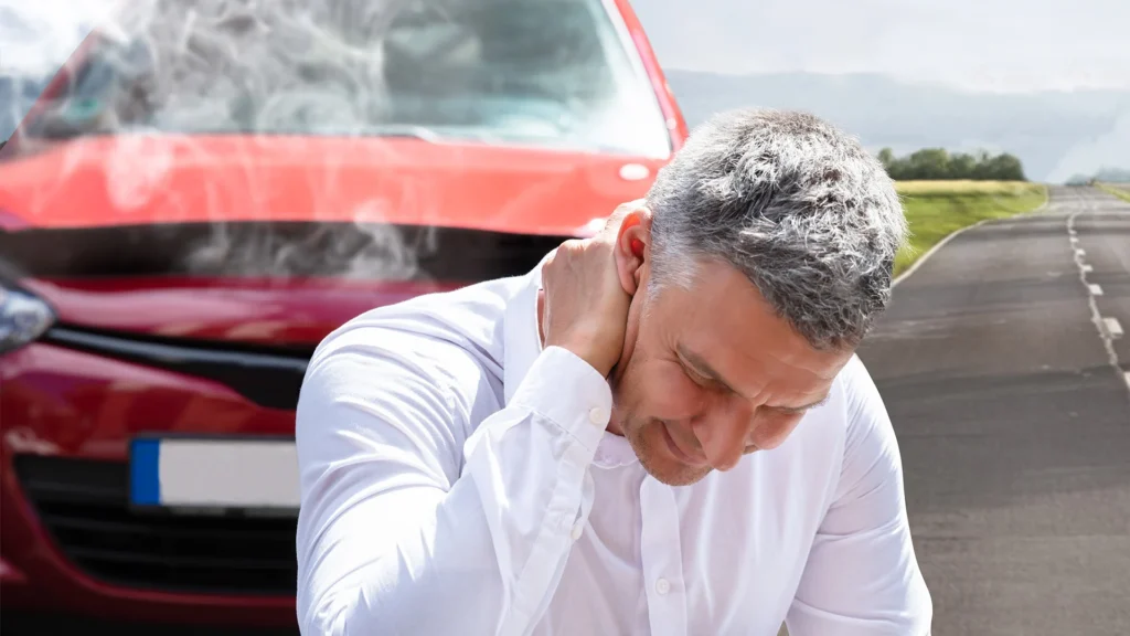 a man holds the back of his neck while sitting in front of a crashed car