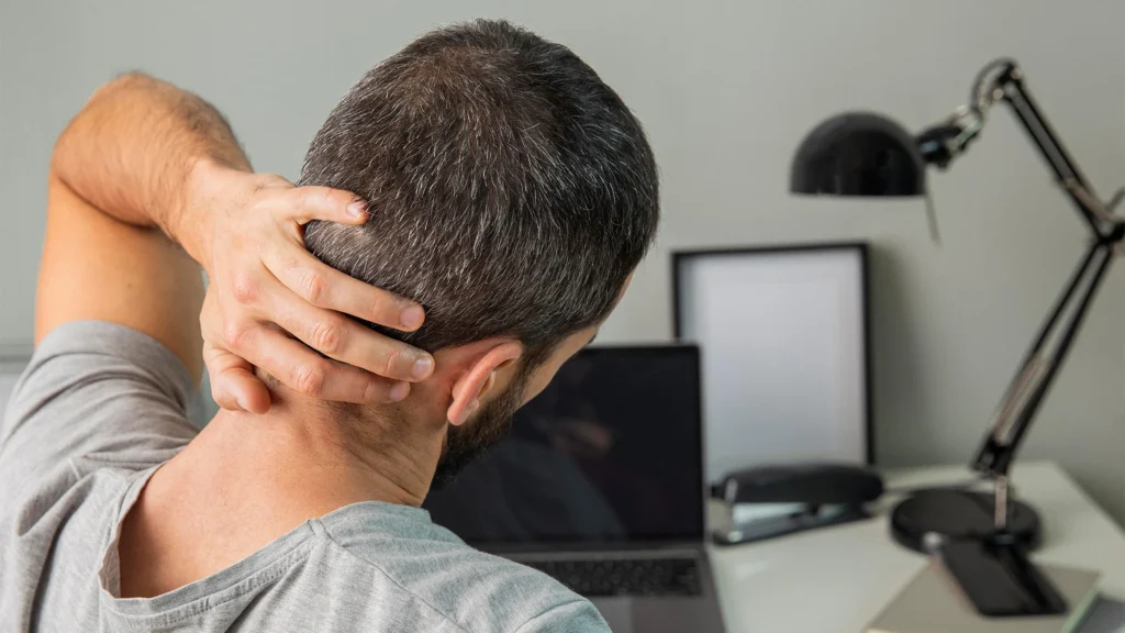 Man With Torticollis At Work Desk Holds The Back Of His Neck