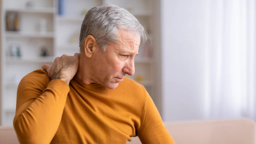 Older Man Sitting On The Couch And Holds His Neck In Pain
