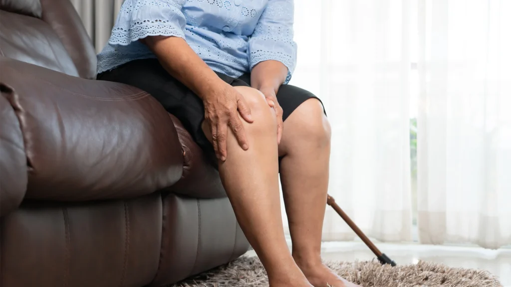Senior Woman Sitting On Couch Holding Knee Pain